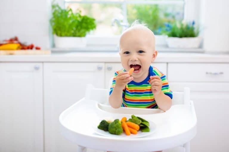 Cute baby eating vegetables in white kitchen. Infant weaning. Little boy trying solid food, organic broccoli, cauliflower, carrot and green peas. Healthy nutrition for kids. Child biting carrot.