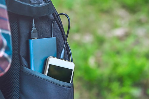 A power bank charges a smartphone in a pocket of a black backpack, on a background of grass