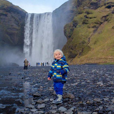 Toddler boy, playing with rocks on the river in fronf of Skogafoss waterfall in Iceland on a sunset cloudy day, autumntime