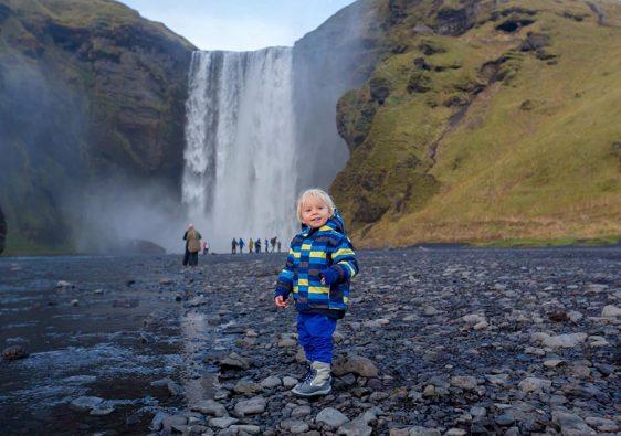 Toddler boy, playing with rocks on the river in fronf of Skogafoss waterfall in Iceland on a sunset cloudy day, autumntime