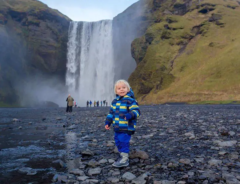 Toddler boy, playing with rocks on the river in fronf of Skogafoss waterfall in Iceland on a sunset cloudy day, autumntime